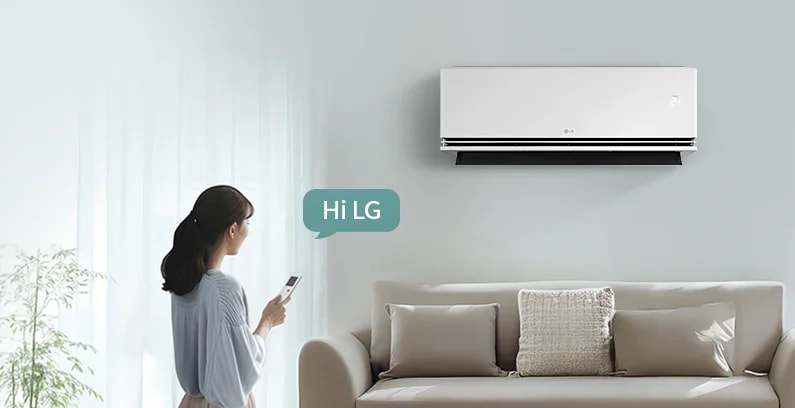 벽에 설치된 LG 휘센 오브제컬렉션 듀얼쿨 벽걸이에어컨이 보입니다. 오른쪽에는 Hi LG라는 말풍선이 있으며, 왼쪽에는 스마트폰을 들고 있는 여성이 서 있습니다. 여성은 창문 쪽을 바라보고 있으며, 창문을 통해 부드러운 자연광이 들어옵니다. 소파는 크림색으로, 여러 개의 쿠션이 놓여 있습니다. 소파 뒤에는 커튼이 드리워져 있고, 왼쪽에는 화분이 놓여 있습니다.