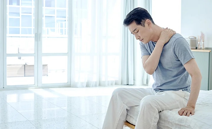 남성이 침대에 앉아 목을 손으로 잡고 있으며, 창문을 통해 밝은 햇살이 들어오는 실내 공간입니다. 남성은 회색 티셔츠와 흰색 바지를 입고 있습니다.