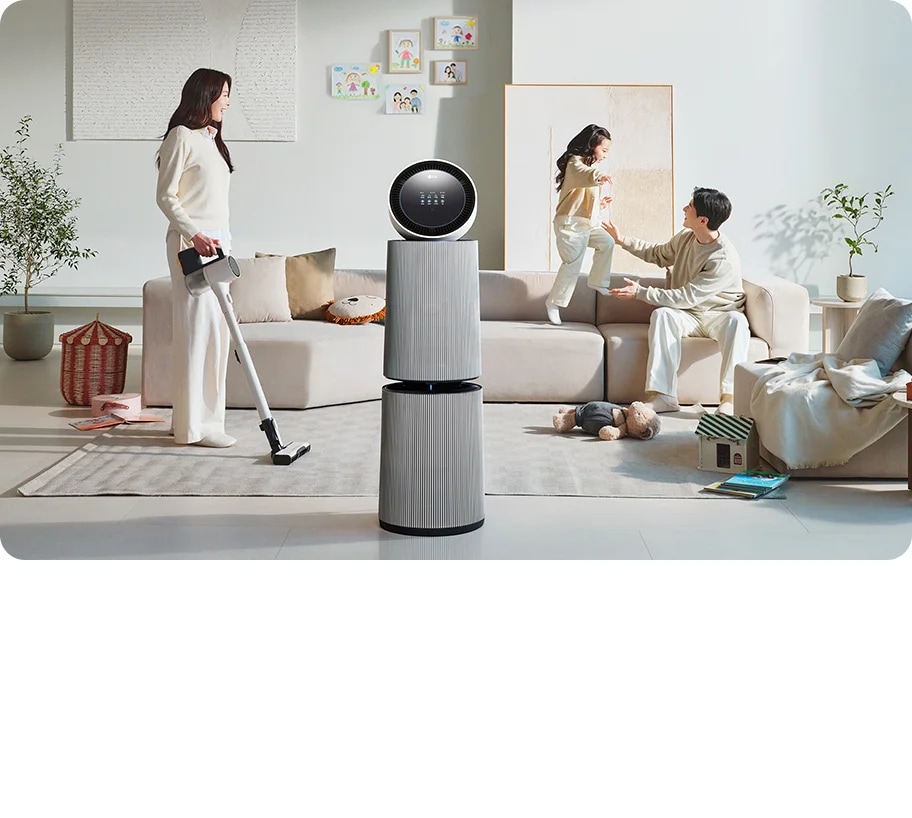 거실 공간에 LG 퓨리케어 AI 오브제컬렉션 360˚ 공기청정기가 있습니다. 소파에 앉아 있는 남성과 뛰어노는 아이, 그리고 청소기를 들고 있는 여성이 보입니다. 벽에는 그림이 걸려 있고, 바닥에는 화분과 장난감 등이 배치되어 있습니다.