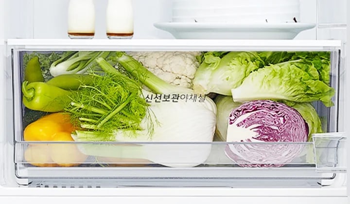 냉장고 내부의 채소 보관 칸에 다양한 채소들이 가득 담겨 있습니다. 노란색과 초록색 피망, 양배추, 브로콜리, 상추, 적양배추 등이 보이며, 채소 칸에는 신선보관야채실이라는 문구가 적혀 있습니다. 위쪽 선반에는 작은 병 두 개와 접시가 놓여 있습니다.