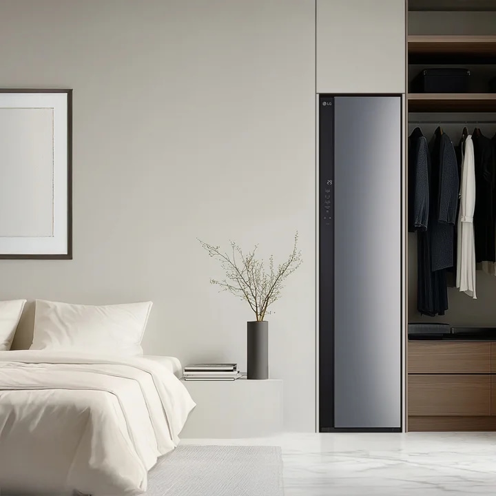 어두운 색상의 LG 스타일러가 설치되어 있으며, 그 옆에는 여러 벌의 옷이 걸려 있습니다. 스타일러 옆에는 나무로 된 서랍과 선반이 있으며, 오른쪽에는 커튼이 드리워져 있고, 왼쪽에는 베이지색 침대와 액자가 걸려 있는 벽이 보입니다. 침대 옆에는 책과 꽃병이 놓여 있습니다.