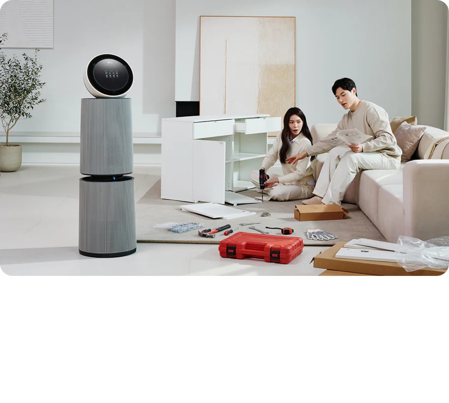 거실 공간에 LG 퓨리케어 AI 오브제컬렉션 360˚ 공기청정기가 놓여 있습니다. 가구를 조립하고 있는 두 사람의 모습이 보입니다. 주변에는 조립 중인 서랍장과 공구가 있습니다.